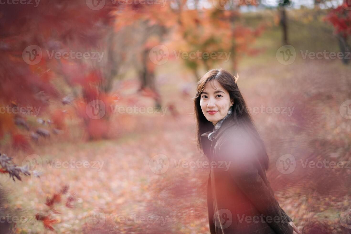 Southeast asian woman smiles gently while standing in serene autumn park surrounded by colorful red and orange leaves creating warm and peaceful atmosphere photo