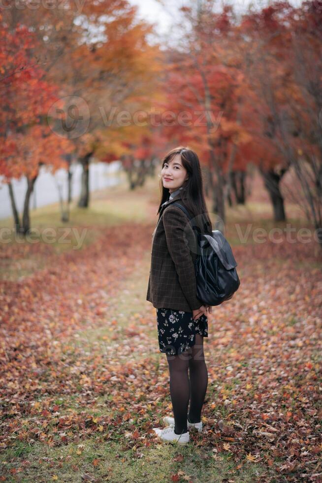 Southeast asian woman stands happily on leaf covered path surrounded by autumn trees wearing brown jacket and black floral skirt with black backpack in serene outdoor setting photo