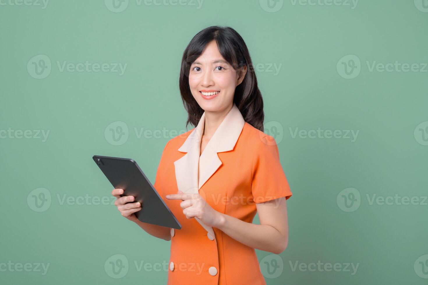 Southeast asian woman in orange suit smiling and holding tablet against green background, showing confidence and modern technology use in professional setting photo