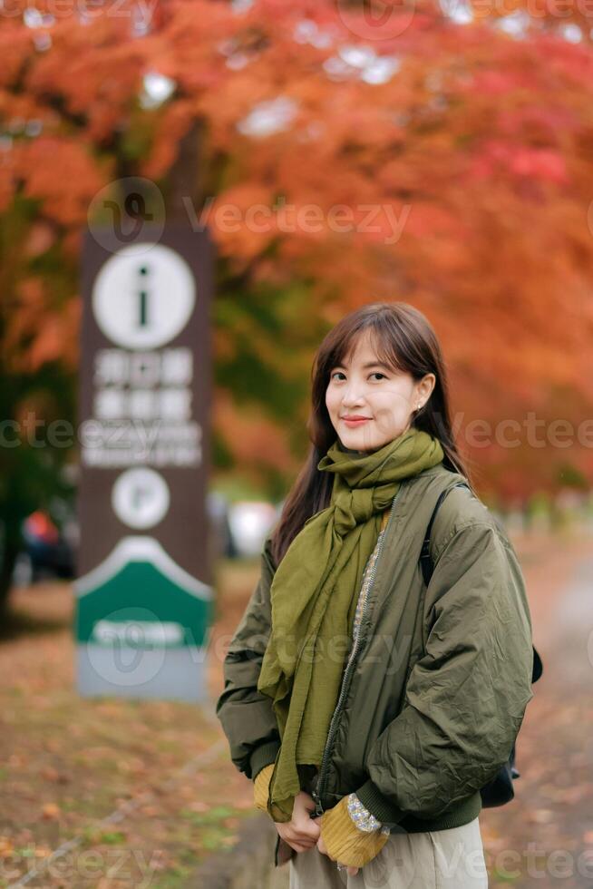 Southeast asian woman stands outdoors wearing green scarf and jacket with gentle smile surrounded by autumn leaves creating warm and peaceful atmosphere in park photo