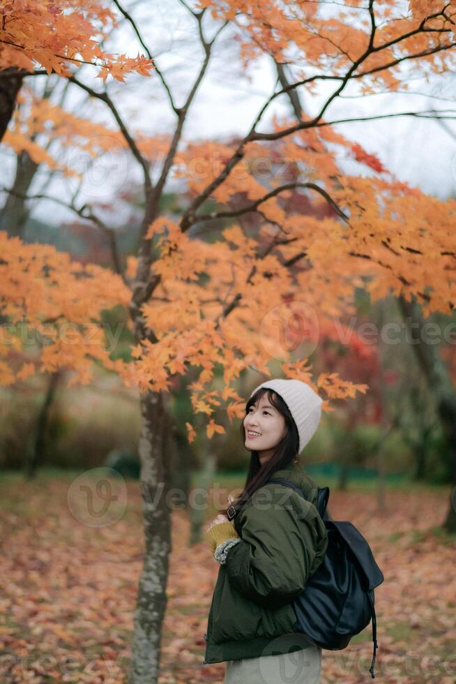 Southeast asian woman enjoys peaceful autumn park wearing warm jacket and hat surrounded by orange leaves and trees creating calm and beautiful seasonal atmosphere photo