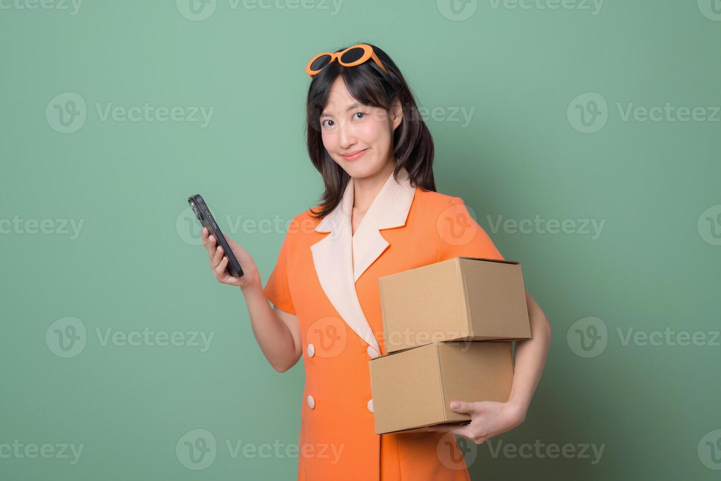 Southeast asian woman in orange dress holding cardboard boxes and using smartphone for internet online shopping with happy expression on green background photo