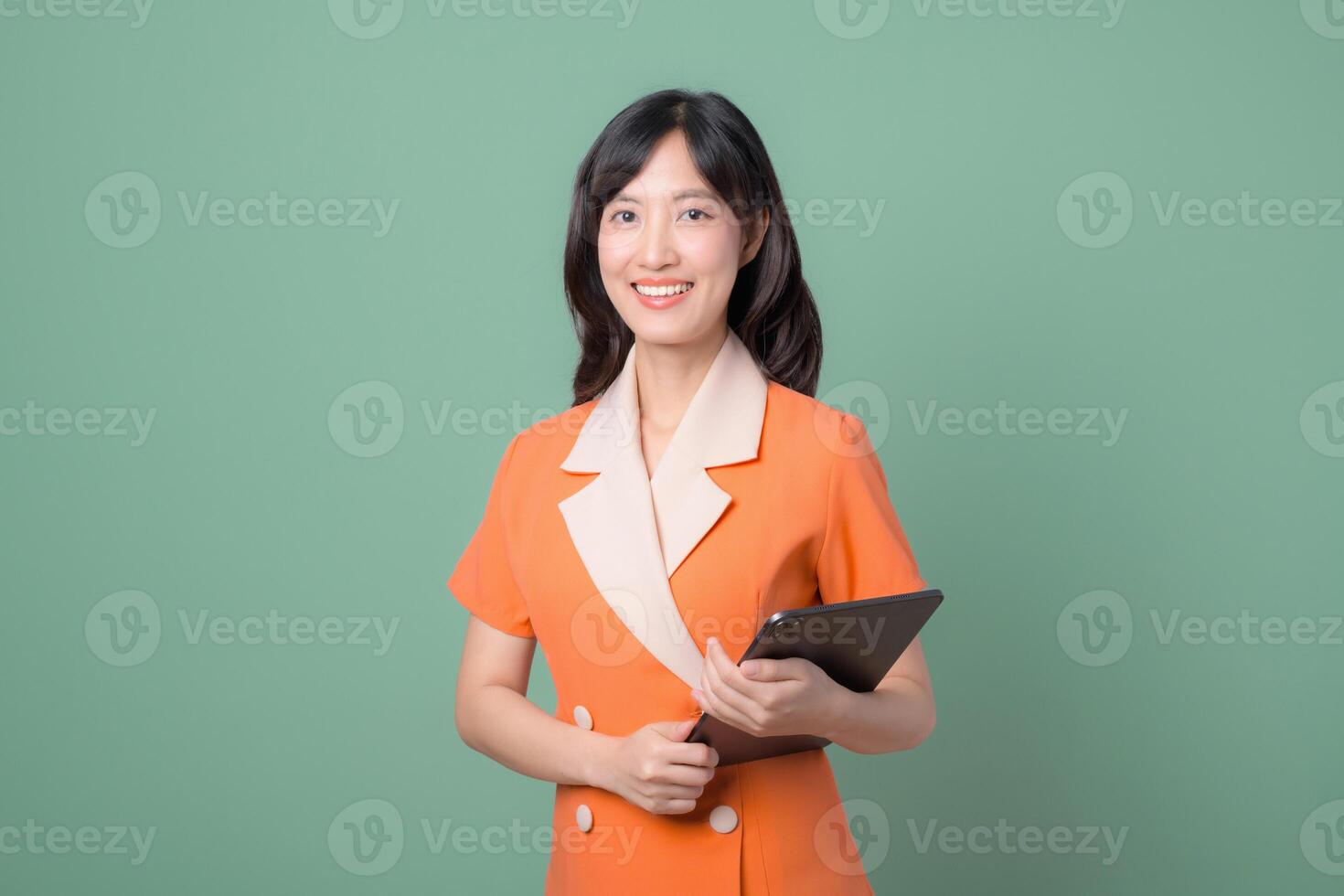 Southeast asian woman in orange dress holding tablet smiling confidently against green background, representing modern professional and positive attitude in business or education photo