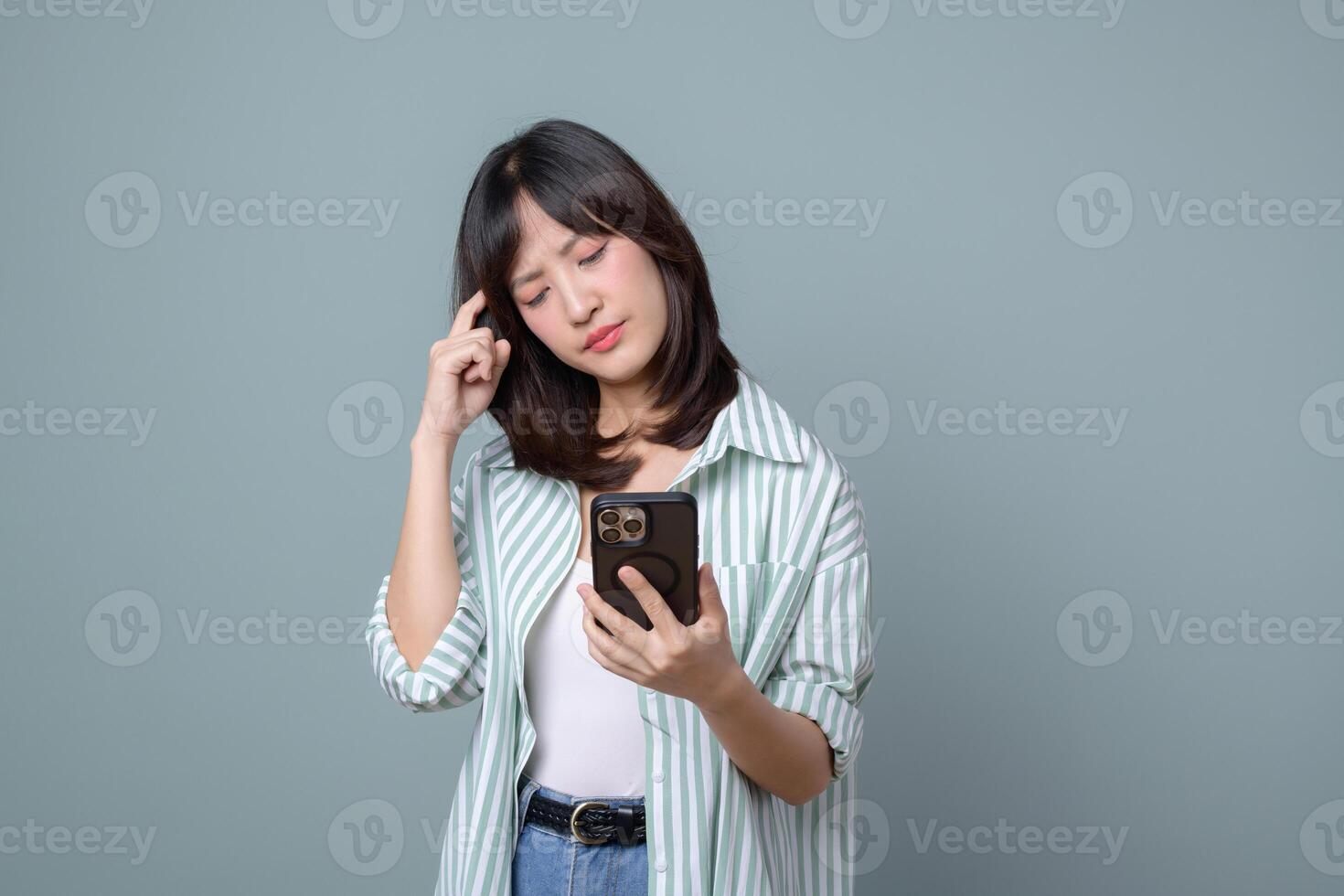 Southeast Asian woman using internet on smartphone looking confused while standing against plain background thoughtful expression photo
