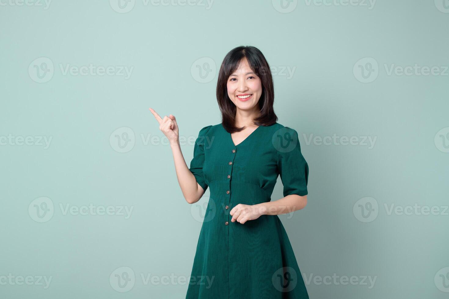 Southeast asian woman in green dress smiling and pointing to side against plain light background, expressing cheerful and positive emotion, suitable for presentation or advertisement concept photo
