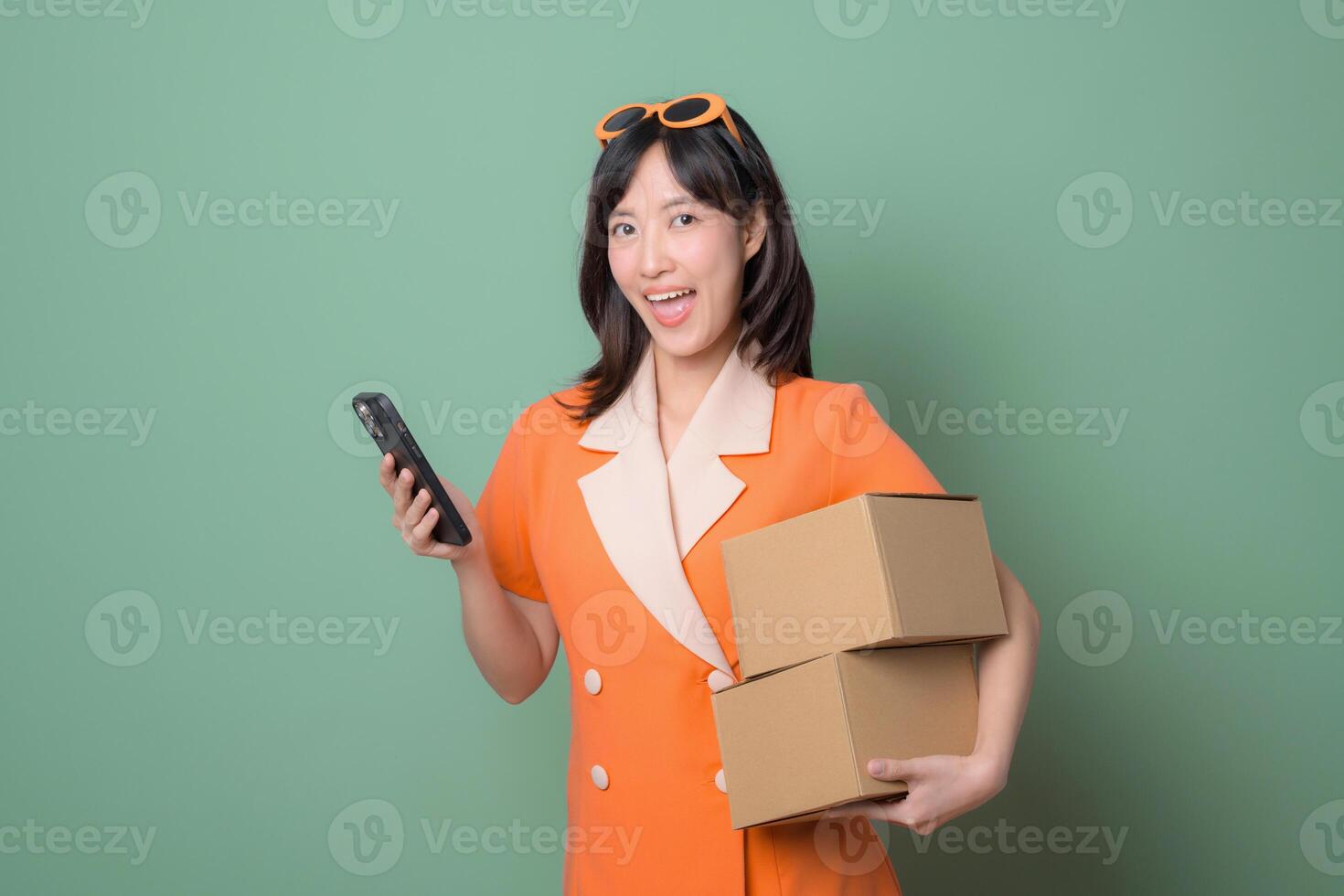 Happy woman in orange dress holds cardboard boxes and phone against green background, delivery and e commerce concept photo
