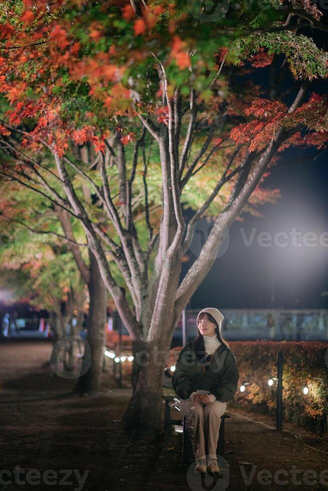 Southeast asian woman sits on bench under colorful autumn trees at night in peaceful park illuminated by soft lights creating calm and serene atmosphere photo