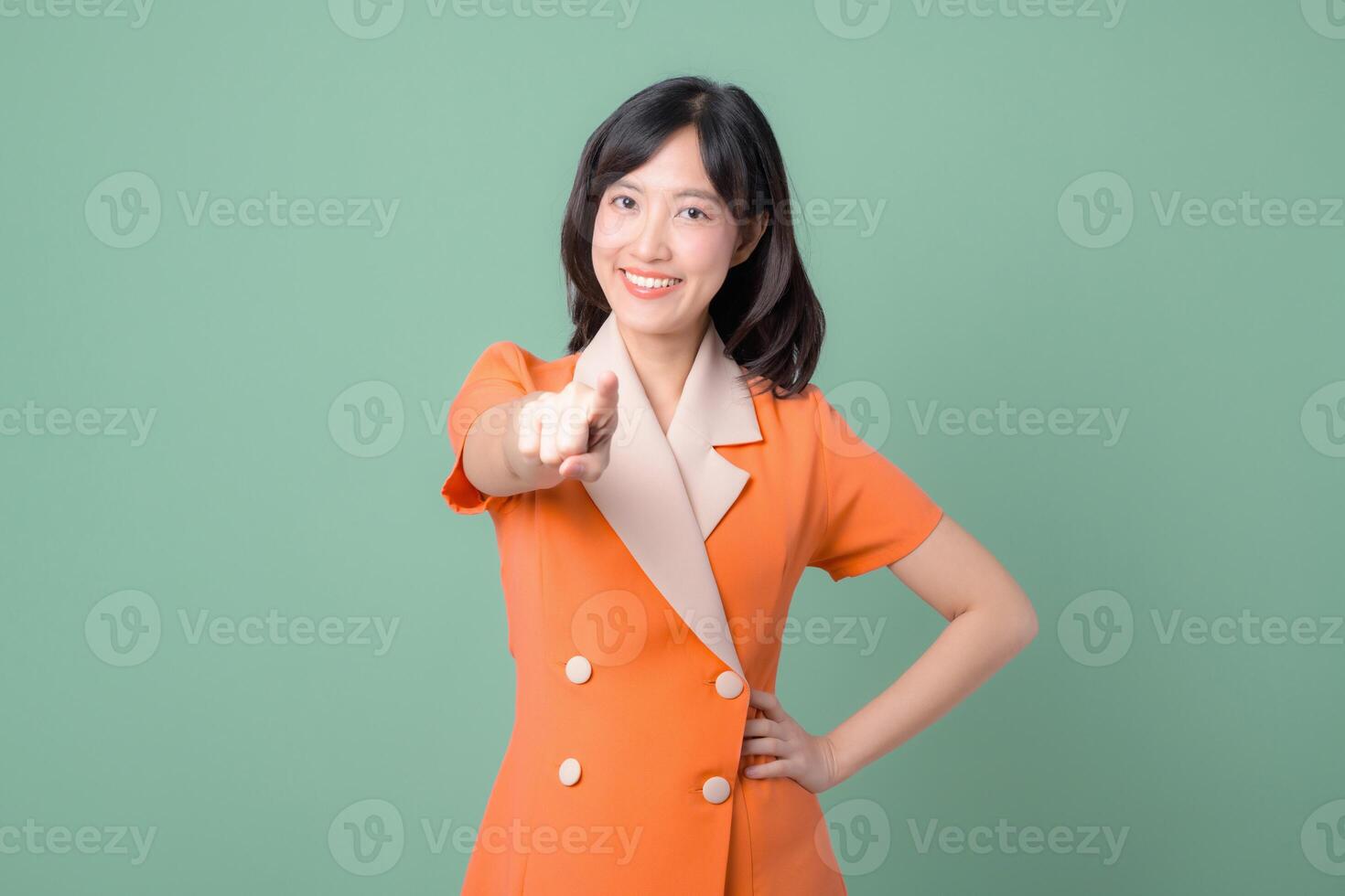 Southeast Asian woman smiling confidently in orange blazer points forward against green background showing positive and friendly expression photo
