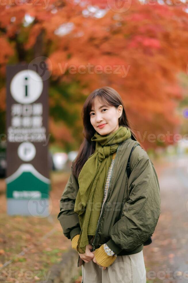 Southeast asian woman wearing green scarf and jacket smiles gently while standing outdoors in autumn with vibrant orange leaves creating warm and peaceful atmosphere photo