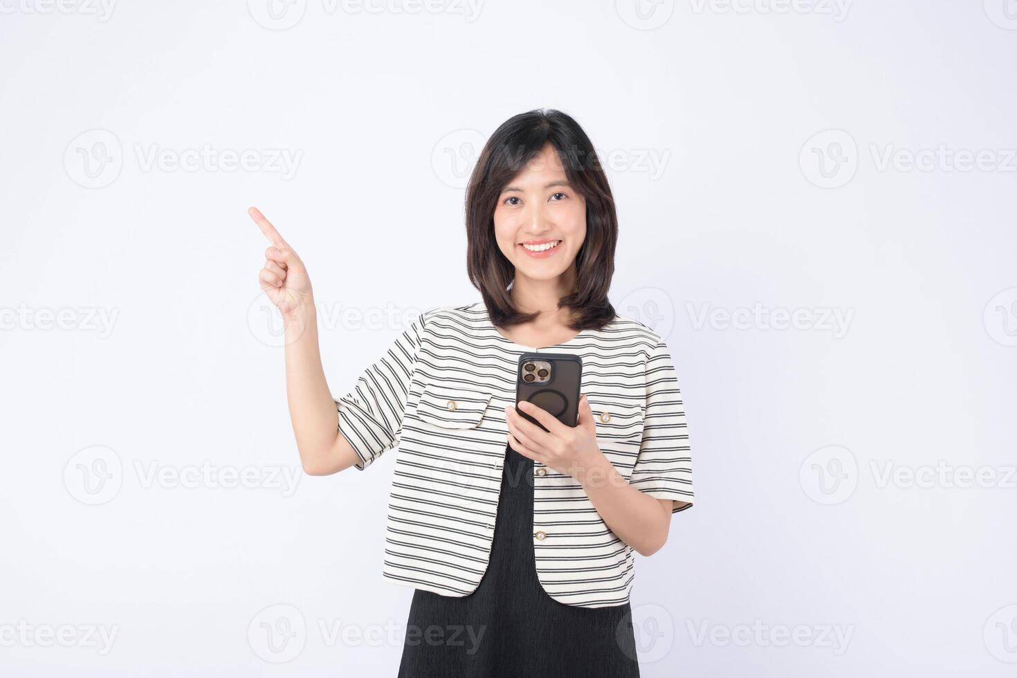 Southeast asian woman smiling while holding mobile phone and pointing up with her finger showing internet application usage in bright studio setting photo