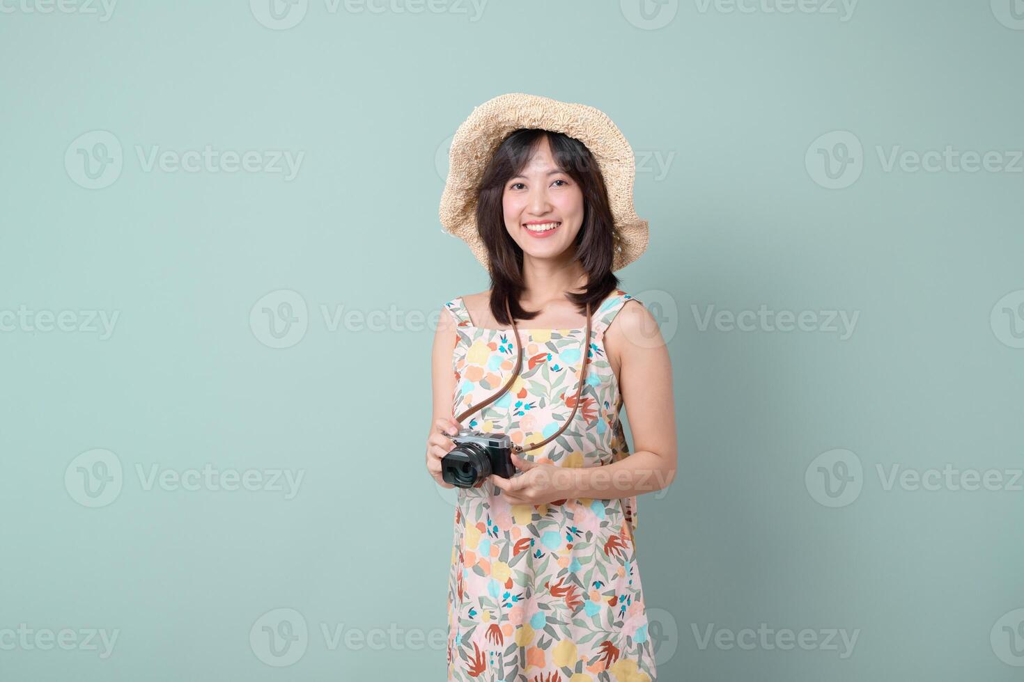 asian woman wearing floral dress and straw hat holds camera smiling happily, capturing travel concept with joy and excitement in simple studio setting photo