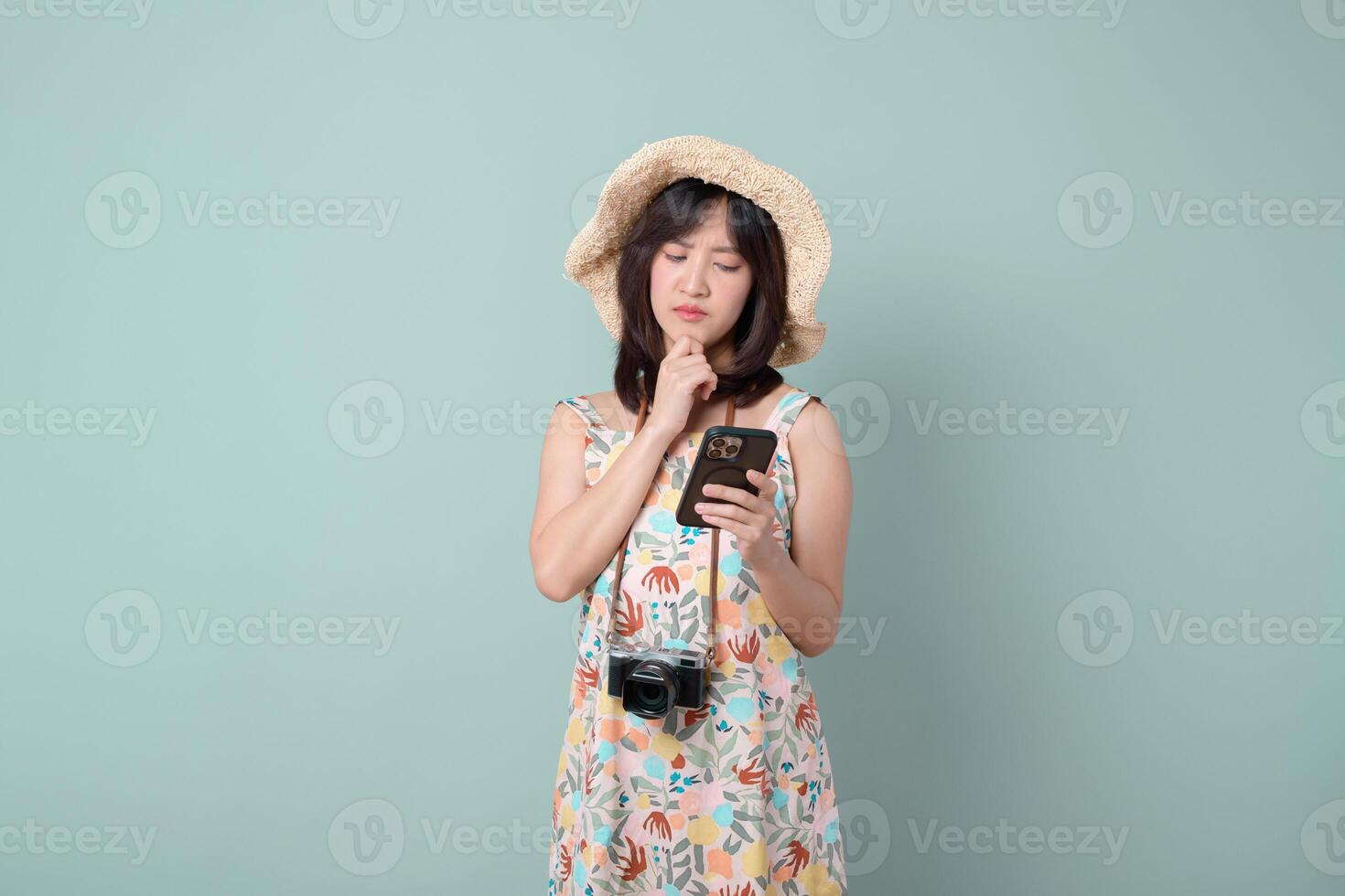 Southeast asian woman wearing hat looks thoughtfully at her mobile phone while holding camera around her neck using internet application for travel planning or communication photo