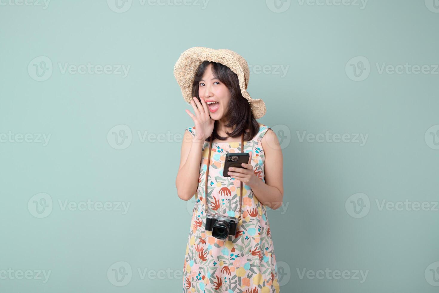 Southeast asian woman happily using mobile phone with internet application while wearing summer hat and floral dress with camera around her neck in studio photo