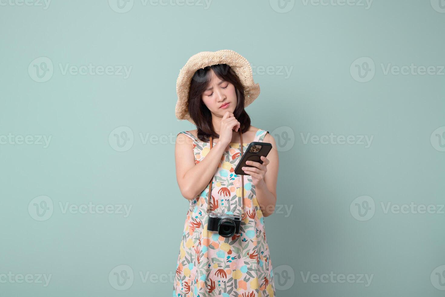 Southeast asian woman wearing hat looks thoughtfully at her mobile phone while holding camera around her neck using internet application for travel or photography photo