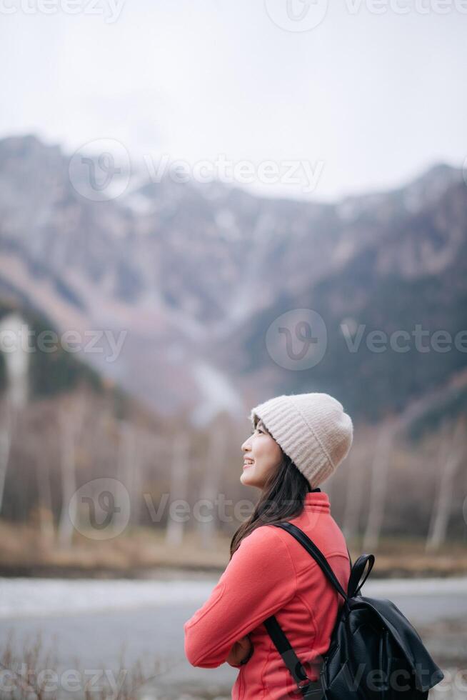 Southeast asian woman wearing warm hat and red jacket enjoys travel concept in mountains with black backpack, smiling peacefully in cold nature scene photo