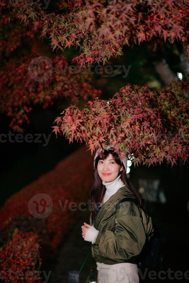 Southeast asian woman travel concept smiling under red autumn leaves at night with casual jacket and backpack photo
