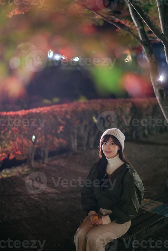 Southeast asian woman enjoys travel concept sitting on bench at night surrounded by autumn leaves and soft glowing lights creating peaceful and warm atmosphere photo