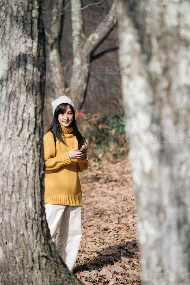 Southeast asian woman wearing yellow sweater and white pants stands among trees in forest during autumn holding phone with calm expression on her face photo