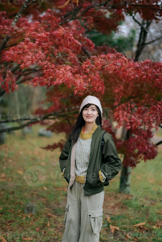 Southeast asian woman stands smiling under vibrant red autumn leaves wearing warm jacket and beanie, enjoying peaceful outdoor fall season with natural beauty around her photo
