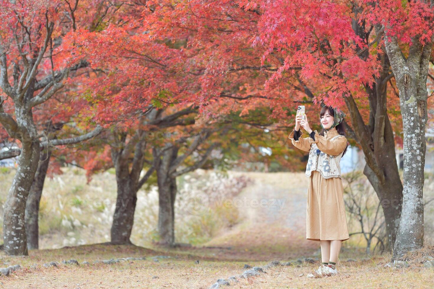 Southeast asian woman enjoys travel concept by taking photo under vibrant red autumn trees in peaceful outdoor setting with joyful expression on her face