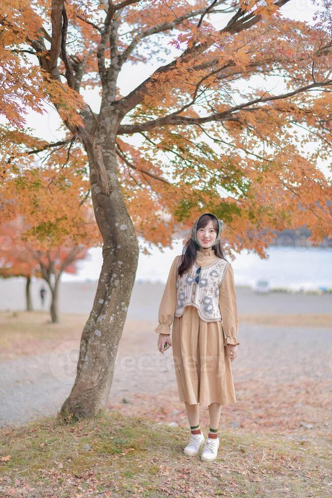 Southeast asian woman enjoys travel concept standing under tree with orange autumn leaves wearing beige dress and headphones in peaceful outdoor setting photo