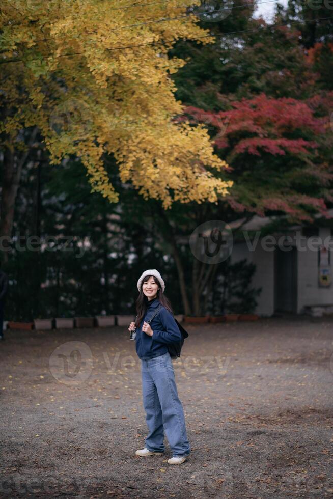Southeast asian woman wearing hat and casual clothes stands smiling under yellow autumn leaves with backpack in peaceful outdoor setting during fall season photo