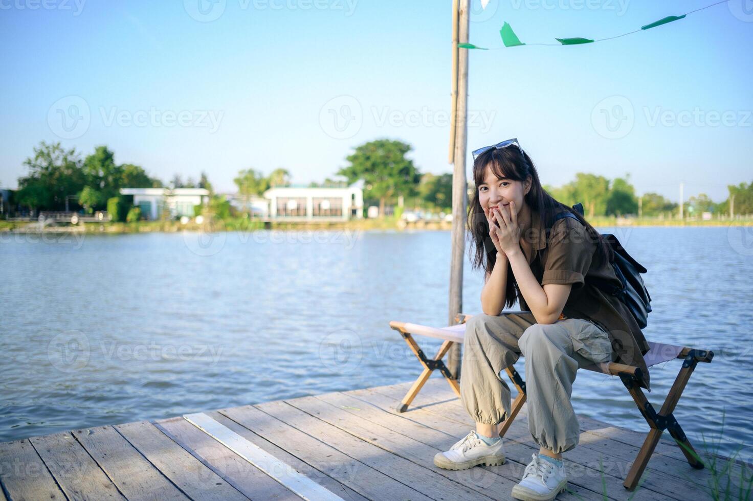 Southeast asian woman enjoys travel concept by sitting on wooden dock near lake with joyful expression and casual outfit on sunny day outdoors photo