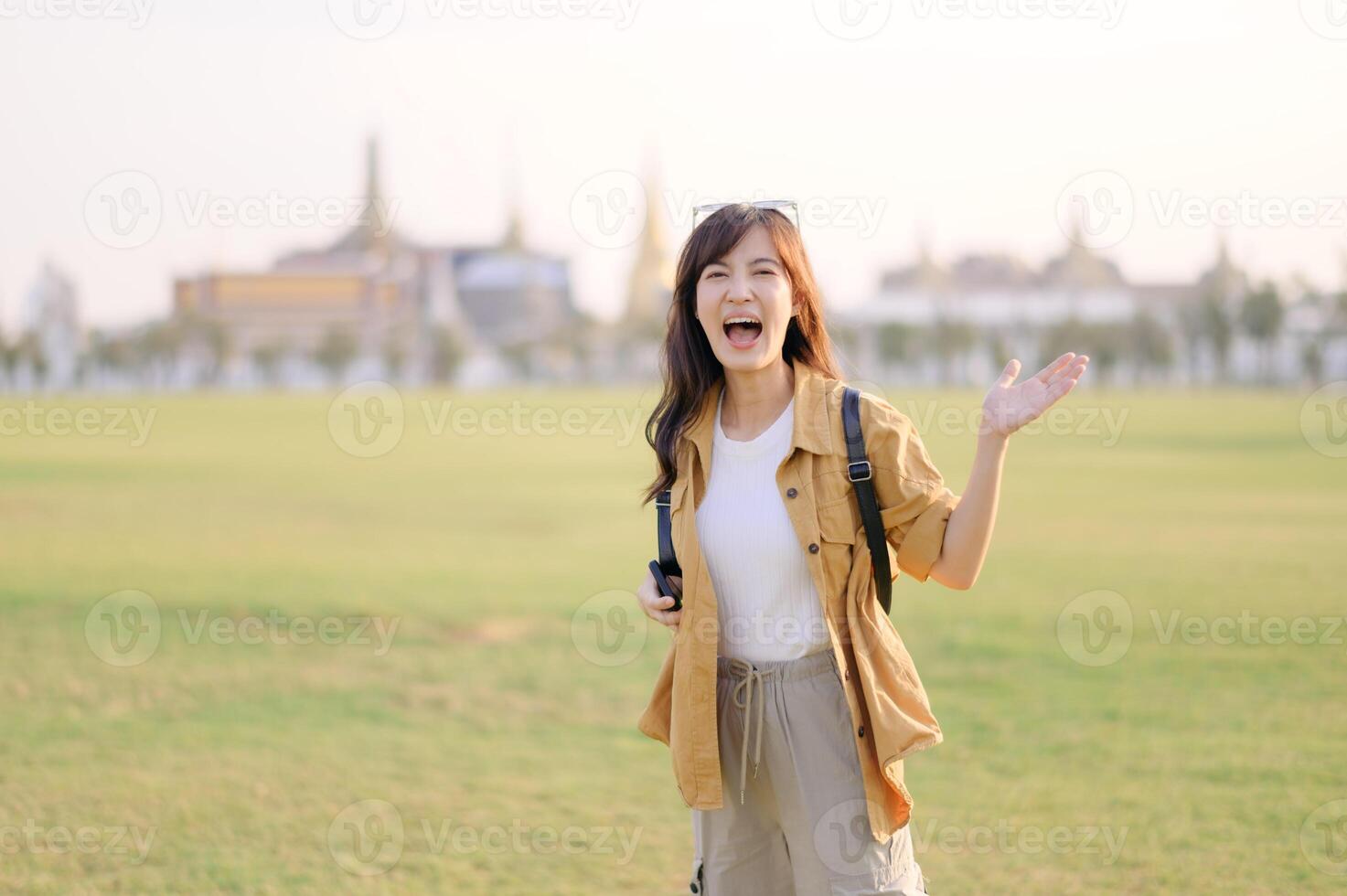 Southeast asian woman travel concept with joyful expression stands in open field waving happily with blurred cityscape in background during bright daylight photo