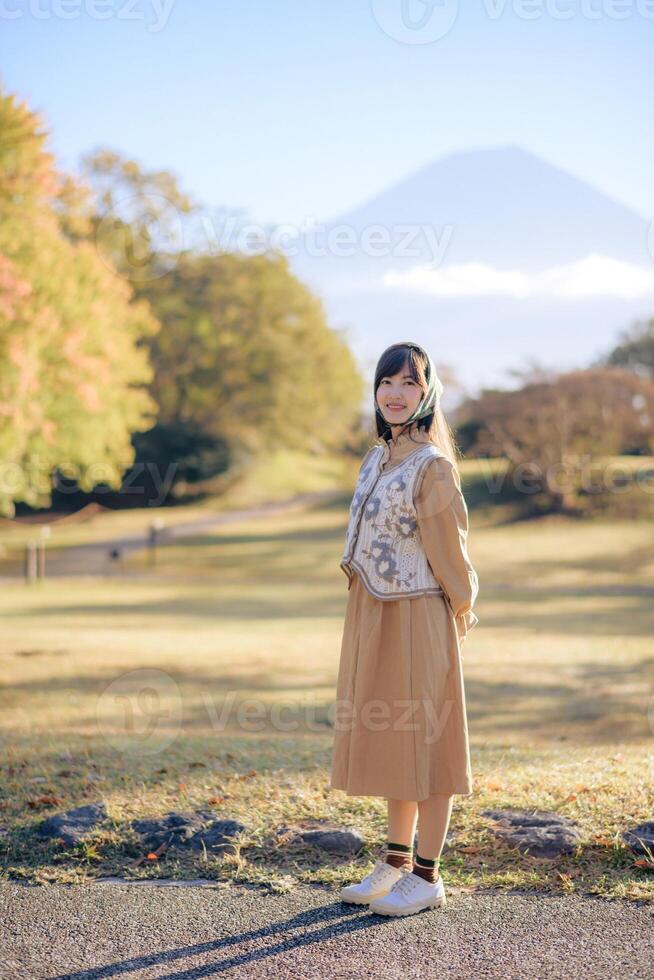 Southeast asian woman stands happily in sunny park wearing beige dress and patterned vest with headscarf, surrounded by autumn trees and distant mountain under clear sky photo
