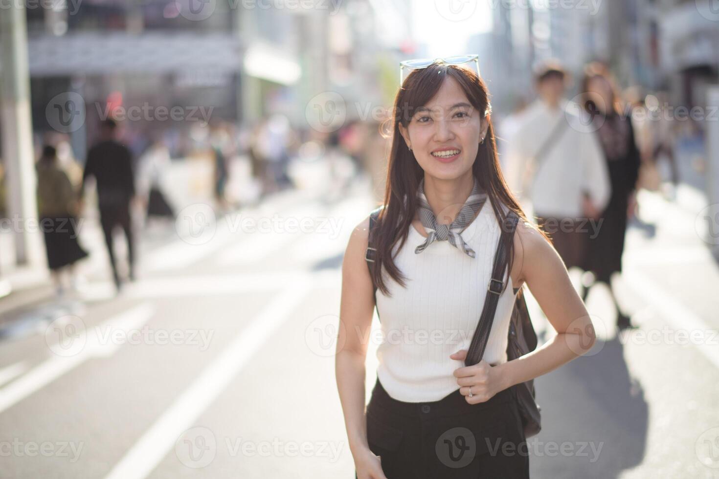 Southeast asian woman smiles warmly while standing on busy city street wearing white sleeveless top and black pants with scarf around her neck on sunny day photo