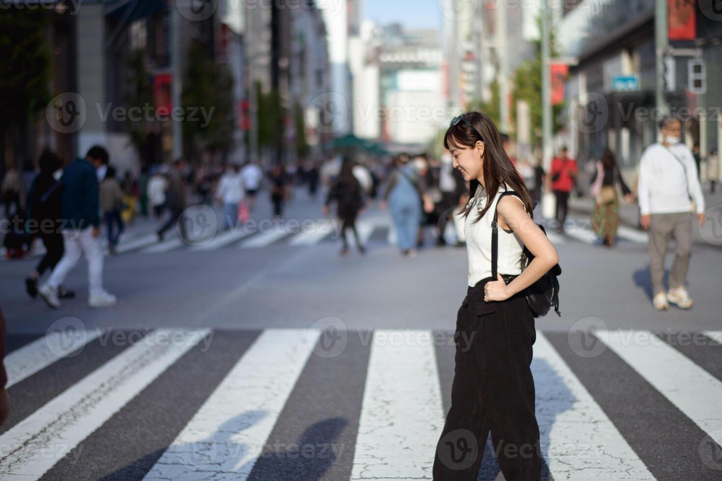 Southeast asian woman walks confidently across busy city street crosswalk on sunny day with smile, surrounded by blurred pedestrians and urban buildings in background photo