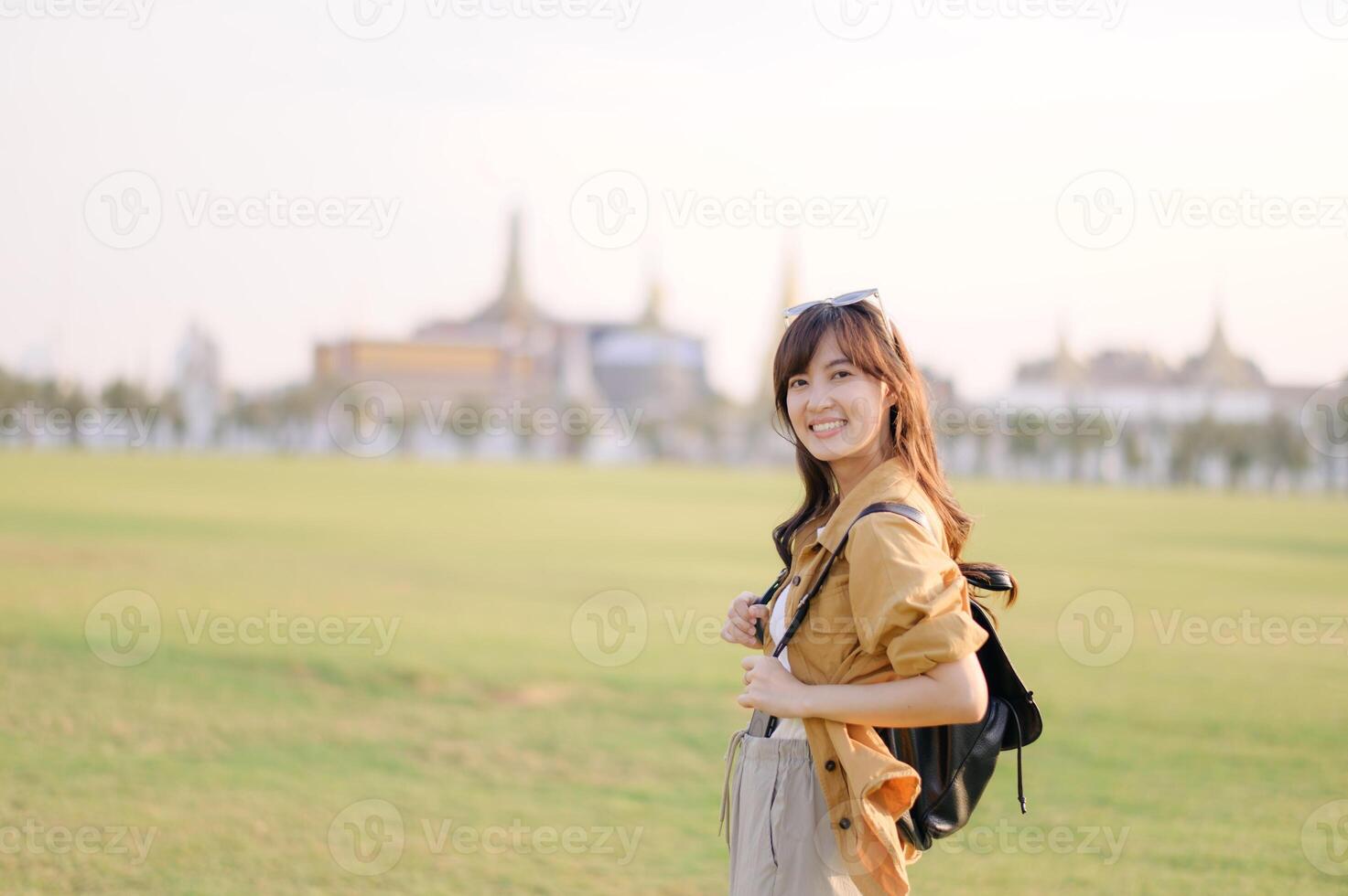 Southeast asian woman smiles happily while travel outdoors with backpack in green field near historic building, capturing joyful travel concept in warm sunlight photo
