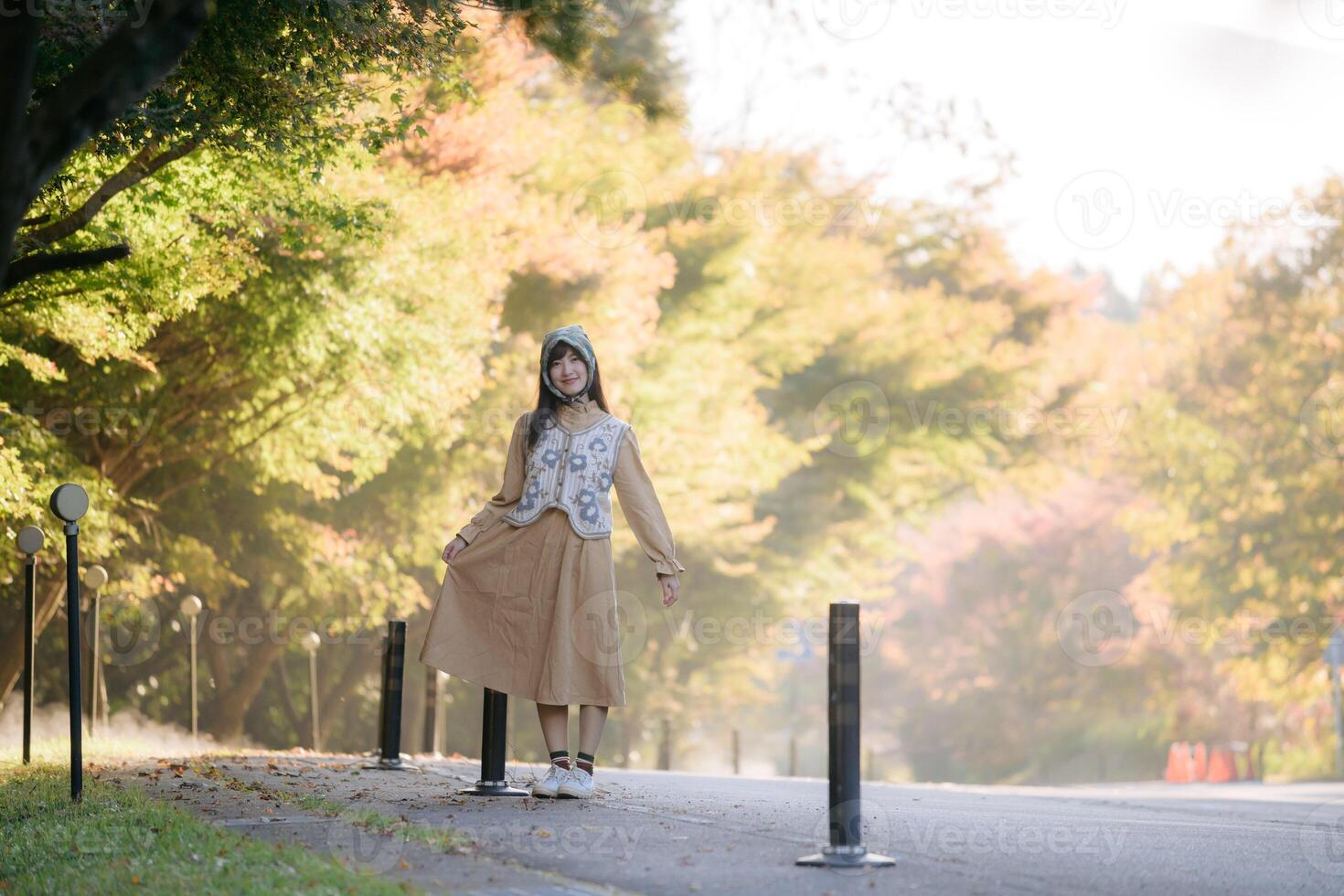Southeast asian woman stands gracefully on tree lined road wearing beige dress and patterned vest surrounded by soft autumn colors and warm natural light photo