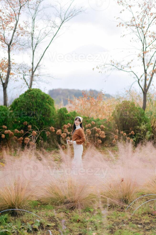 Southeast asian woman stands peacefully in field of soft pink grass surrounded by autumn trees and green bushes under cloudy sky creating calm and serene atmosphere photo