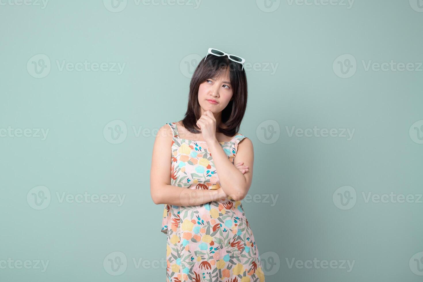 Southeast asian woman in colorful floral dress poses thoughtfully for advertising against plain light green background with sunglasses on her head photo