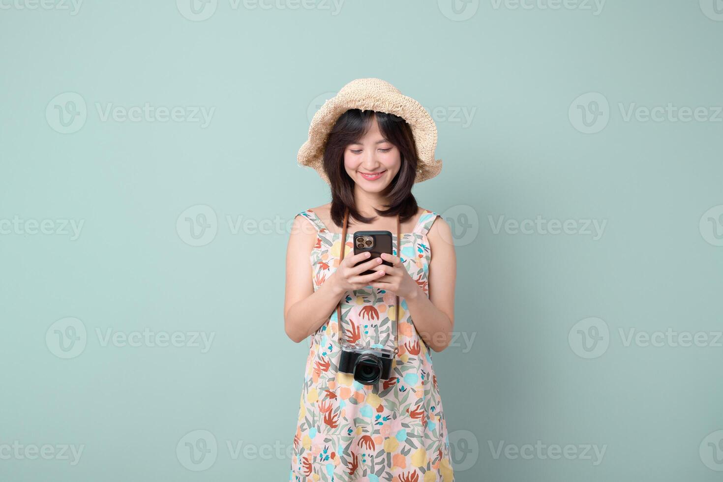 Southeast asian woman wearing floral dress and straw hat happily uses mobile phone to access internet application while camera hangs around her neck in front of plain background photo