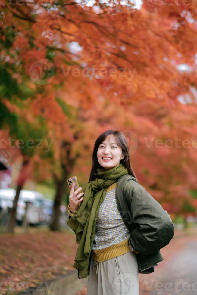 Southeast asian woman smiles warmly while holding phone outdoors during autumn with vibrant orange leaves in background creating cozy and joyful atmosphere photo