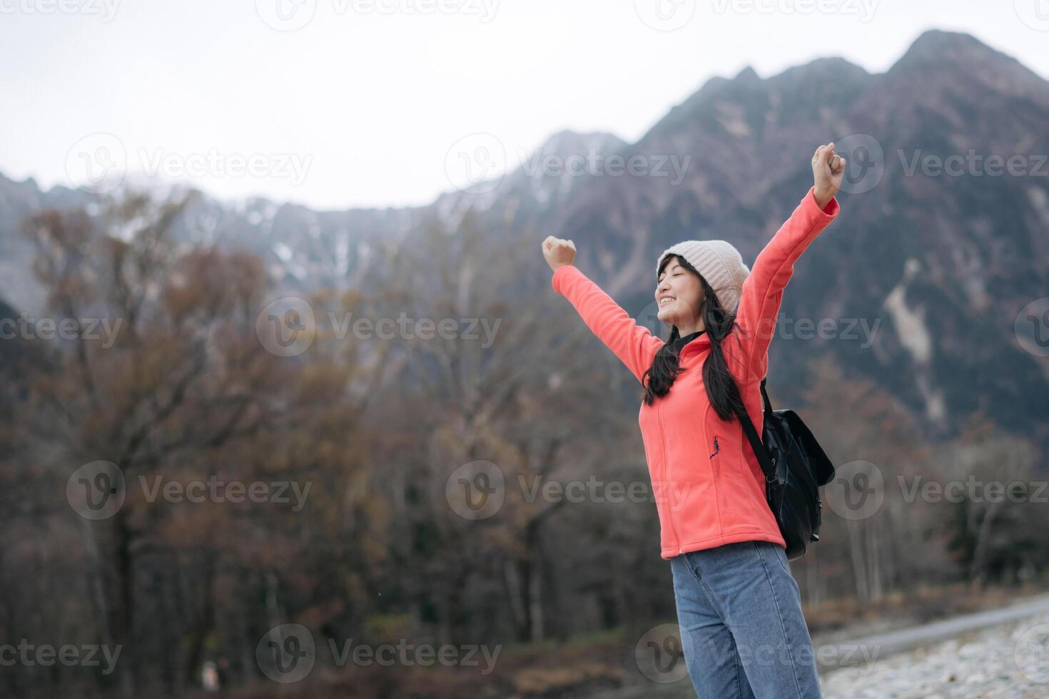 Southeast asian woman travel concept with joyful expression wearing pink jacket and beanie standing outdoors with mountain background and trees in cool weather photo