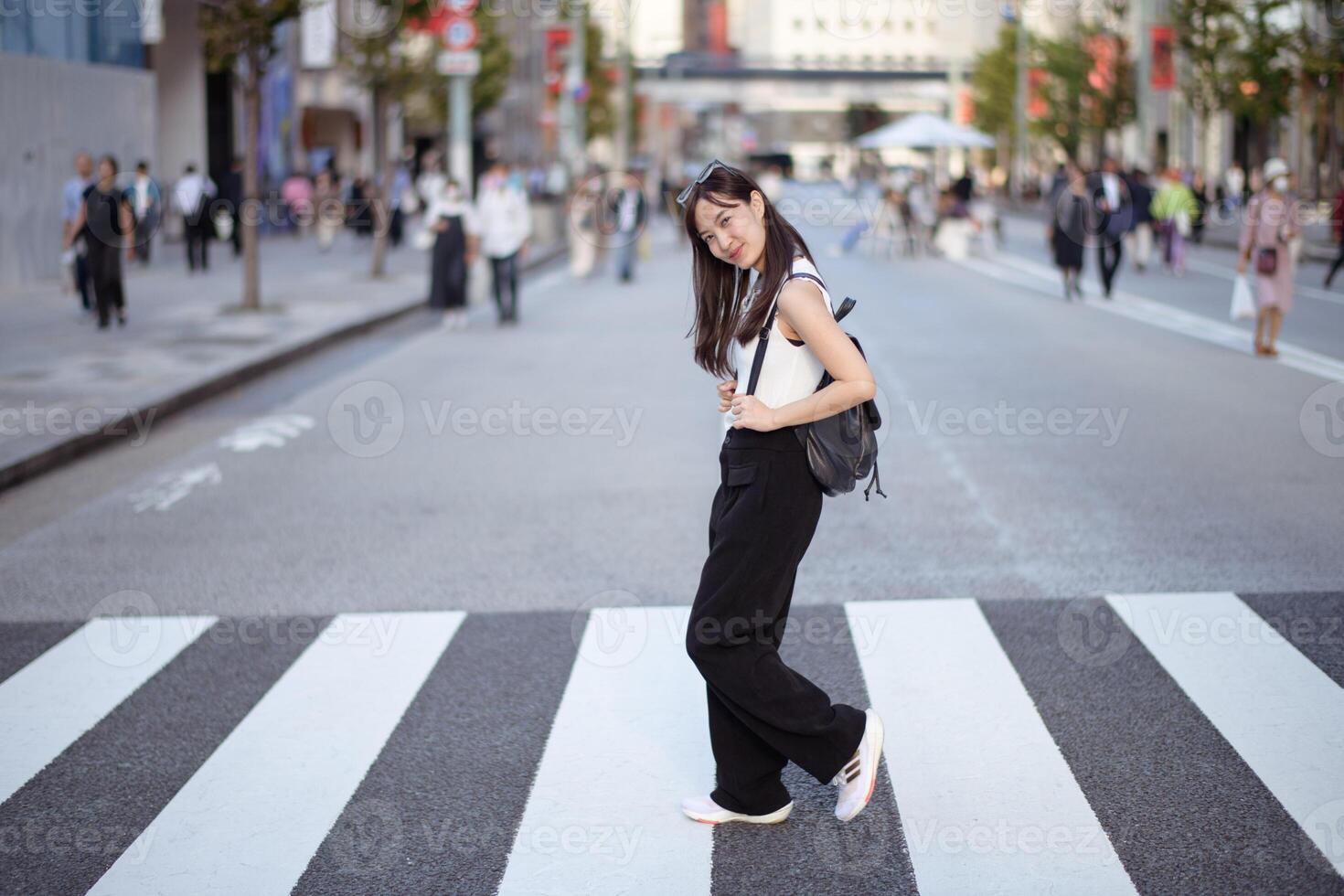 Southeast asian woman confidently walks across city street crosswalk wearing casual black pants and white top with backpack on bright day in urban environment photo