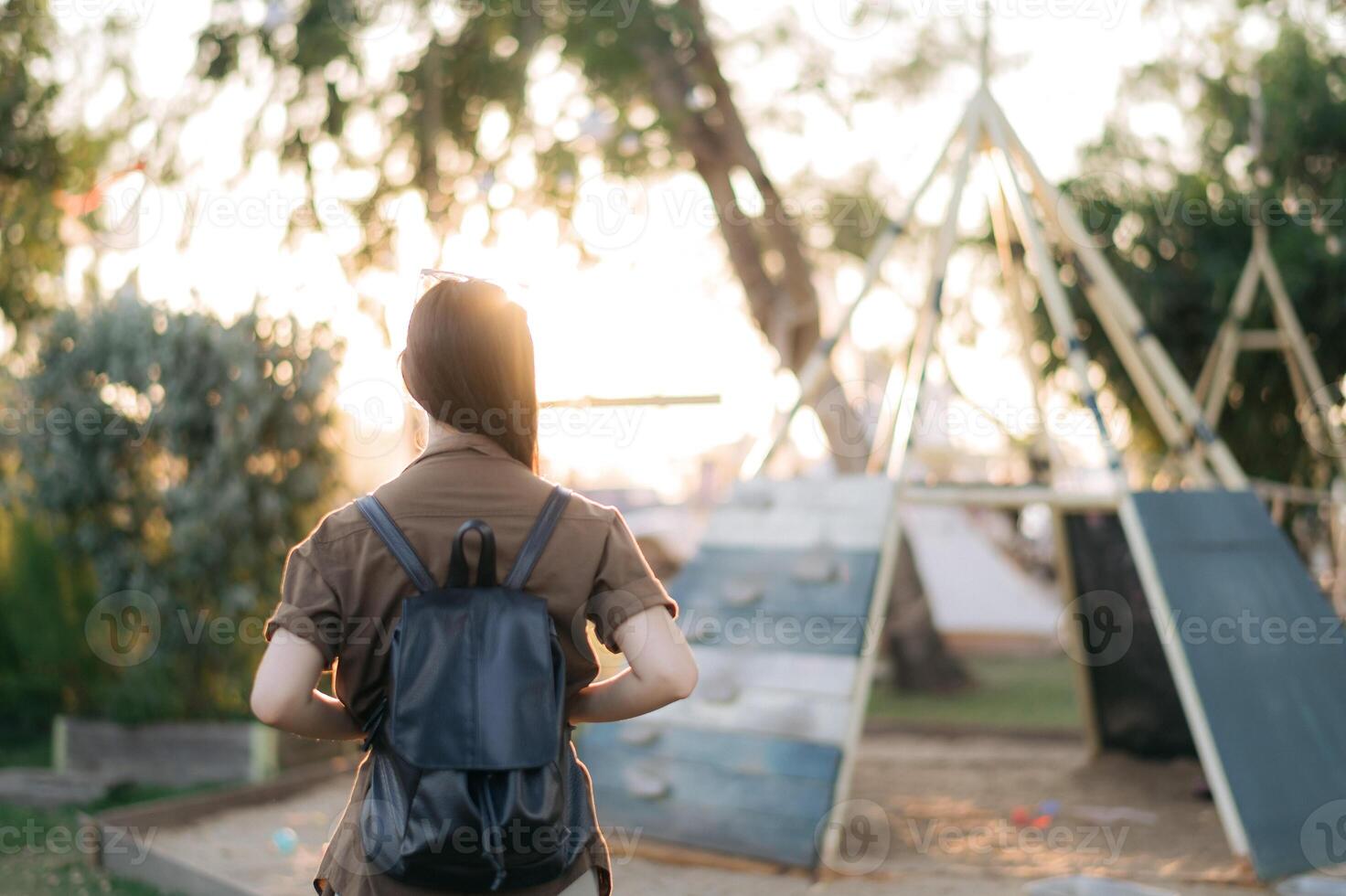 Southeast asian woman with backpack enjoys travel concept outdoors in park during sunset with playground equipment and trees in background creating peaceful mood photo