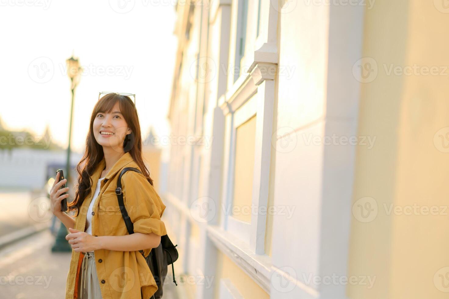 Southeast asian woman enjoys travel concept with bright smile holding phone outdoors near light colored building in warm sunlight creating happy and relaxed mood photo