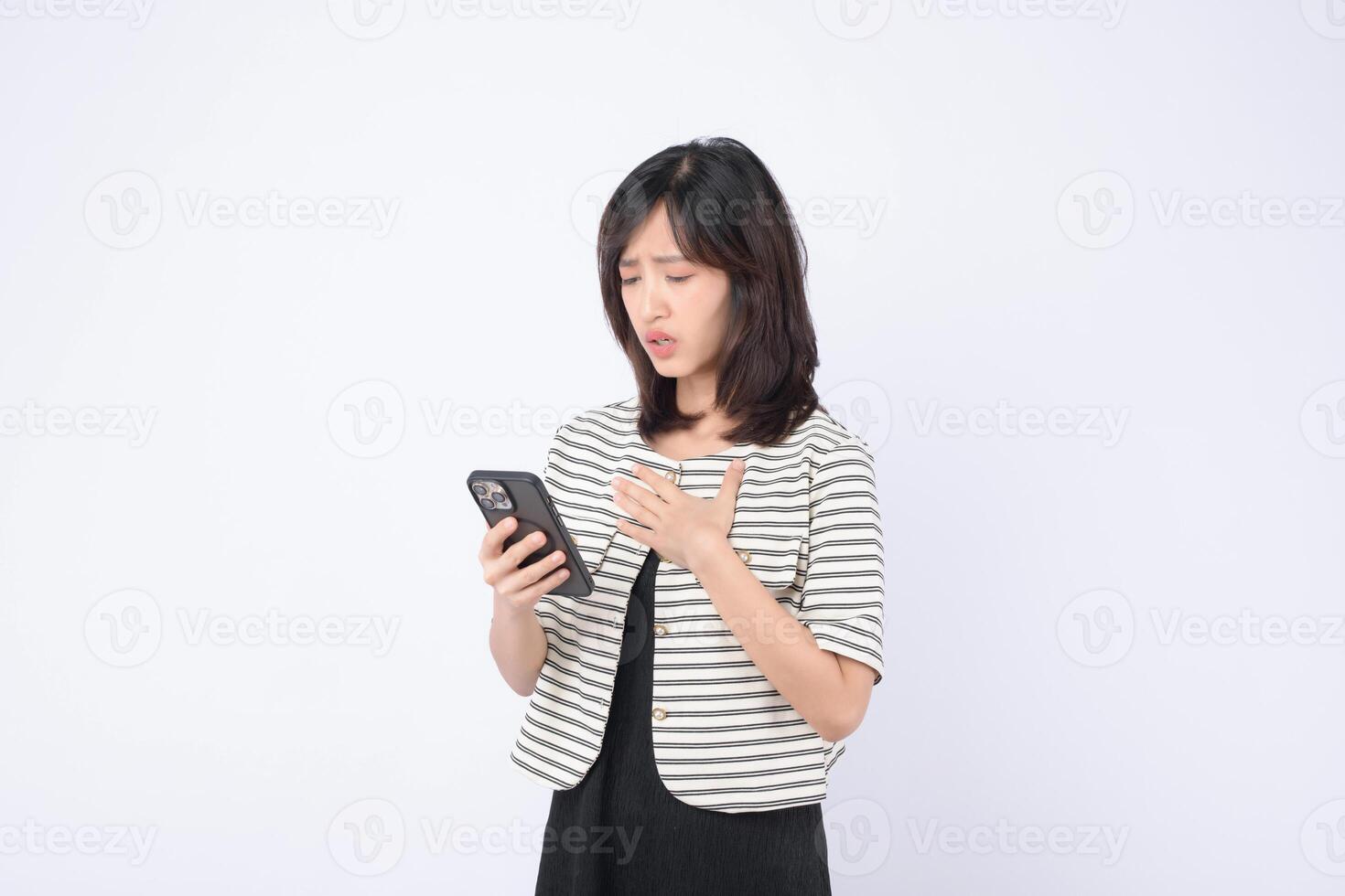 Southeast asian woman in striped jacket holding smartphone with worried expression standing against white background, emotional reaction to message or news photo