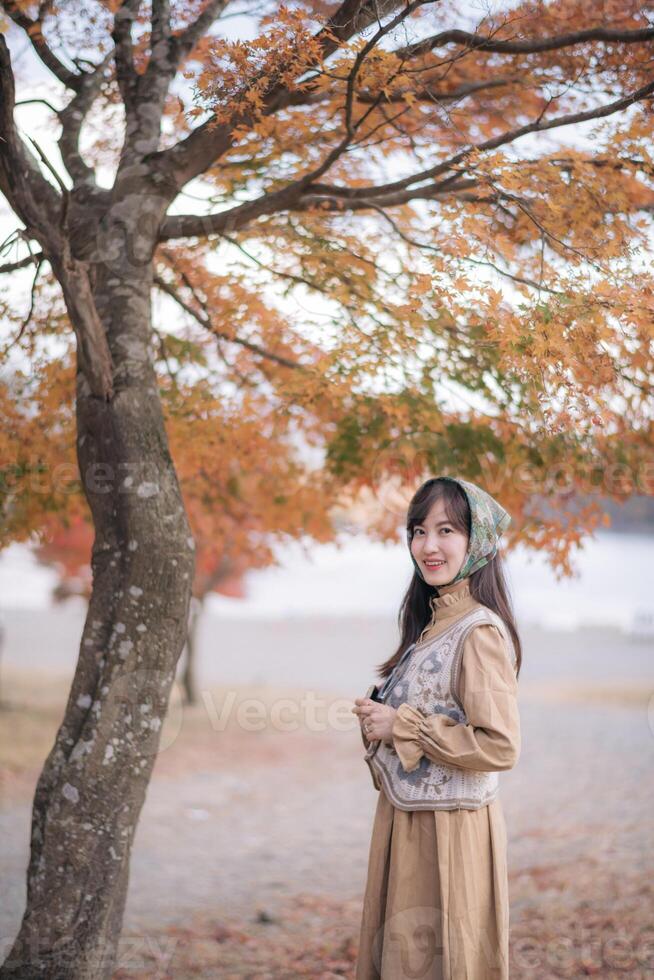 Southeast asian woman enjoys travel concept in autumn park wearing vintage style dress and headscarf smiling under orange leaves tree with peaceful natural background photo