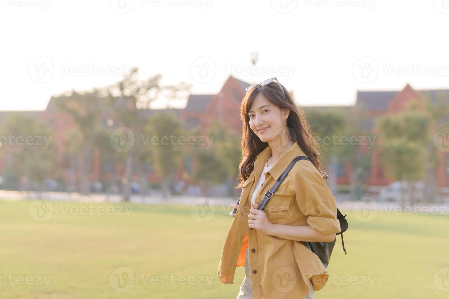 Southeast Asian woman enjoying travel outdoors with cheerful smile, wearing casual clothing and backpack, standing on green grass with sunlight in background photo