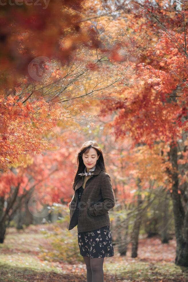 Southeast asian woman stands peacefully among autumn trees with vibrant orange and red leaves wearing dark floral dress and brown jacket enjoying serene fall atmosphere photo