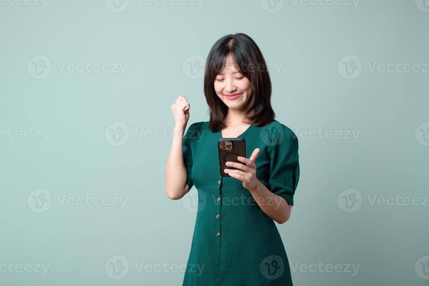 Southeast asian woman happily uses mobile internet application on her phone showing excitement and joy in simple green dress against plain background photo