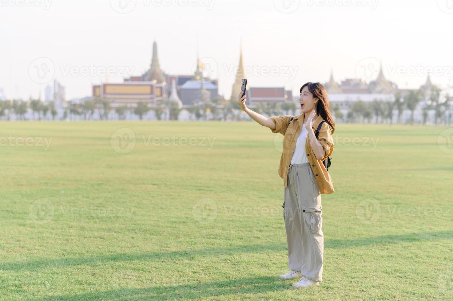 Southeast asian woman enjoys travel concept by taking selfie in large green field with historic temple buildings in background under bright daylight photo