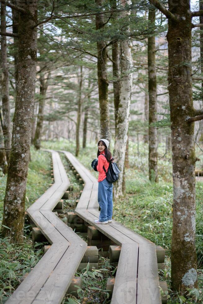Southeast Asian woman enjoys peaceful walk on wooden path through lush forest wearing casual clothes and backpack with joyful expression on her face photo