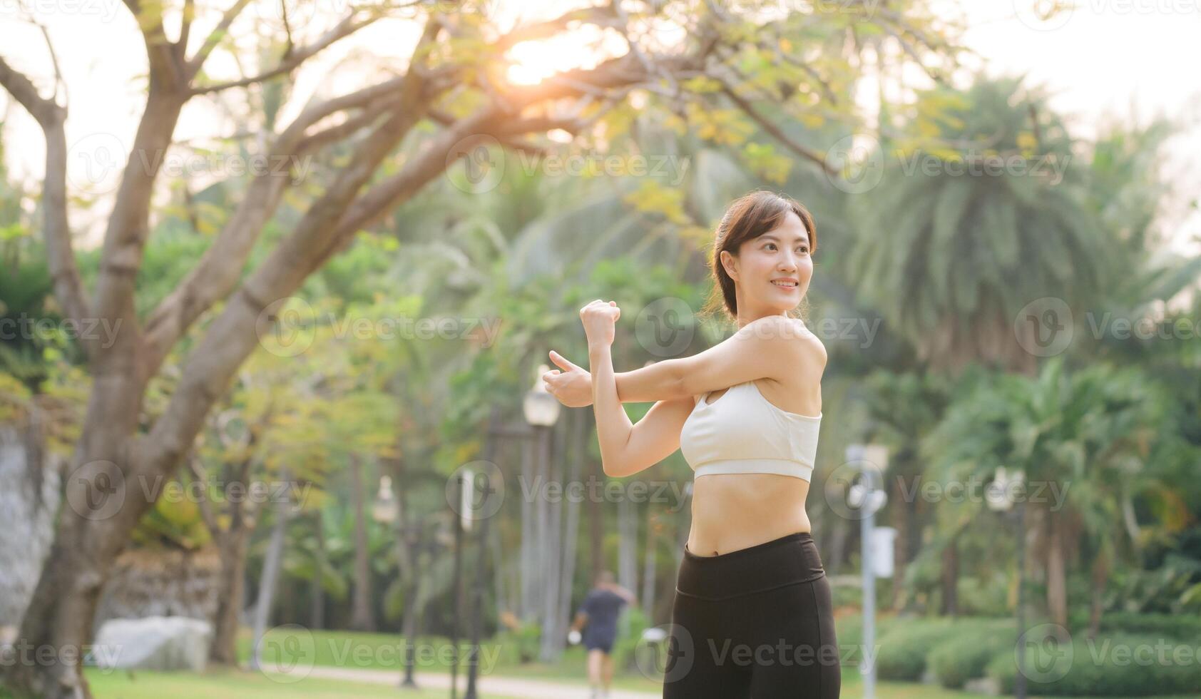 Southeast asian woman enjoying healthy lifestyle concept by doing exercise outdoors in park, stretching arms with happy expression surrounded by green nature and sunlight photo