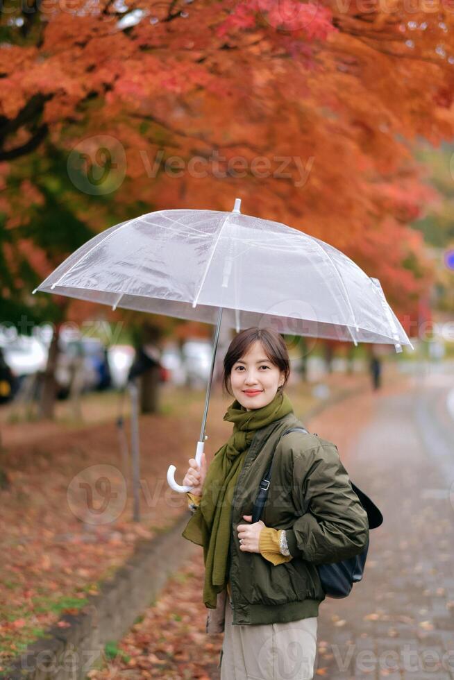 Southeast asian woman stands smiling under transparent umbrella on rainy autumn day with vibrant orange leaves creating warm and peaceful atmosphere in background photo